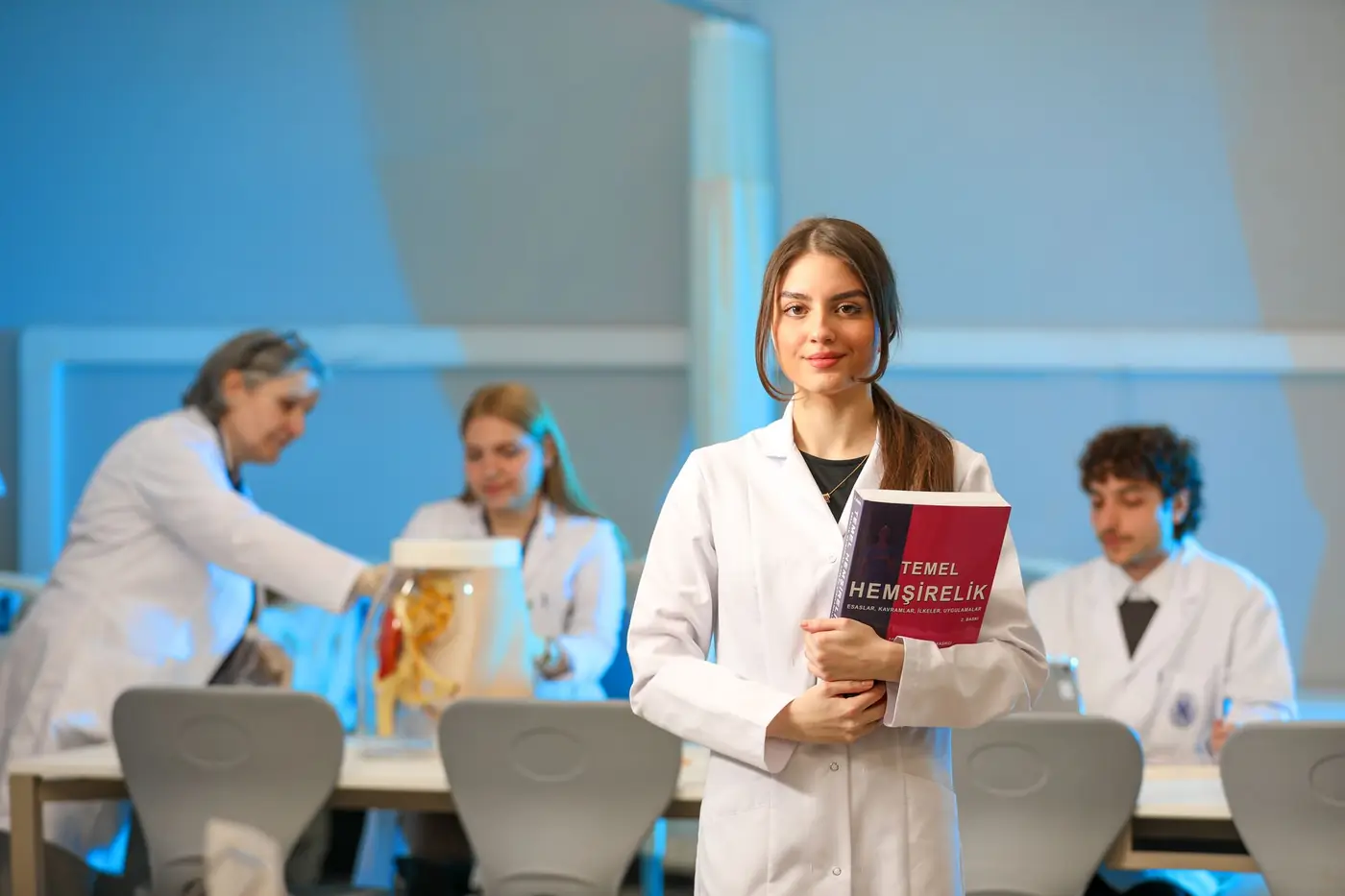 A medical student holding a textbook in an anatomy laboratory, with classmates working at a table behind.