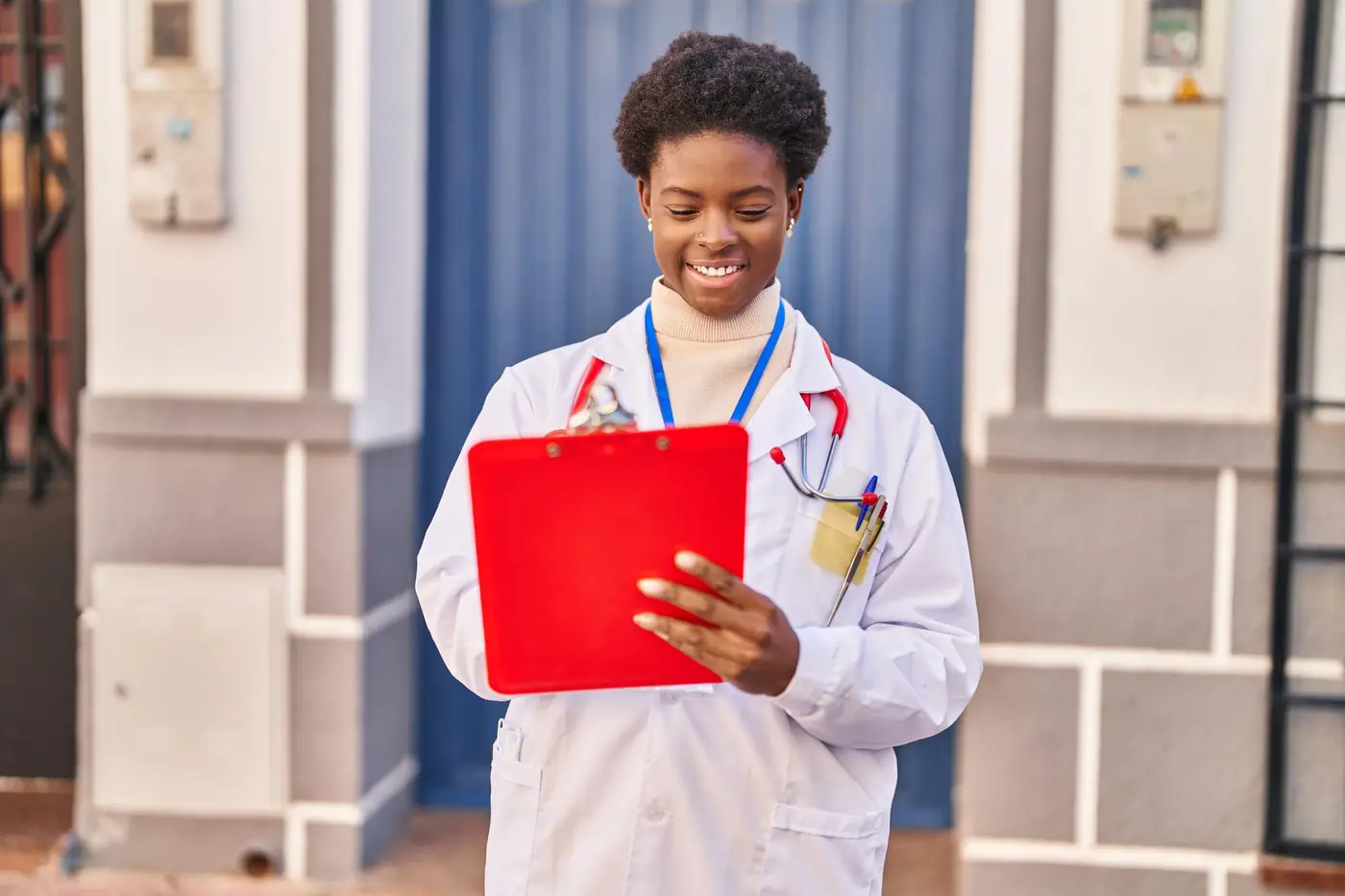 A medical student in a white coat smiling while reading a clipboard outdoors.
