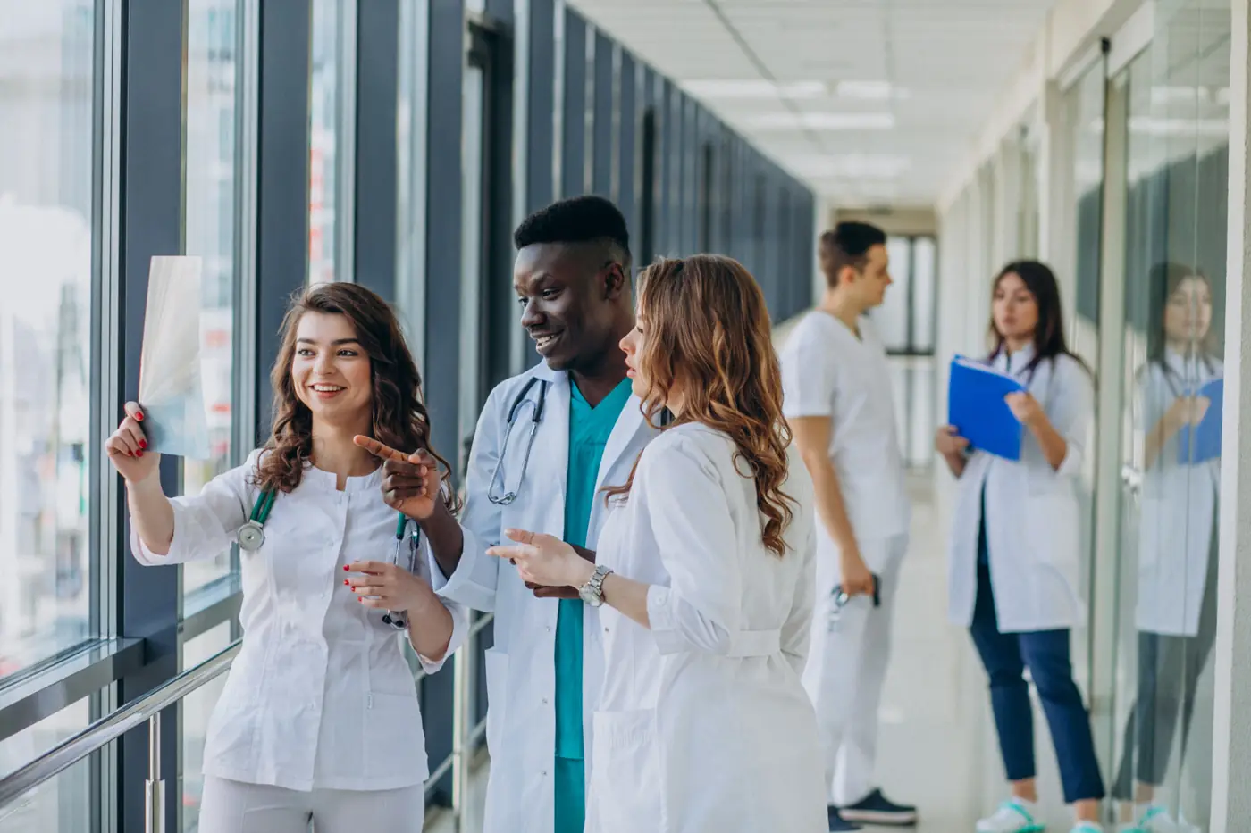 Three medical students at a window examining an X-ray film together while colleagues talk behind them.