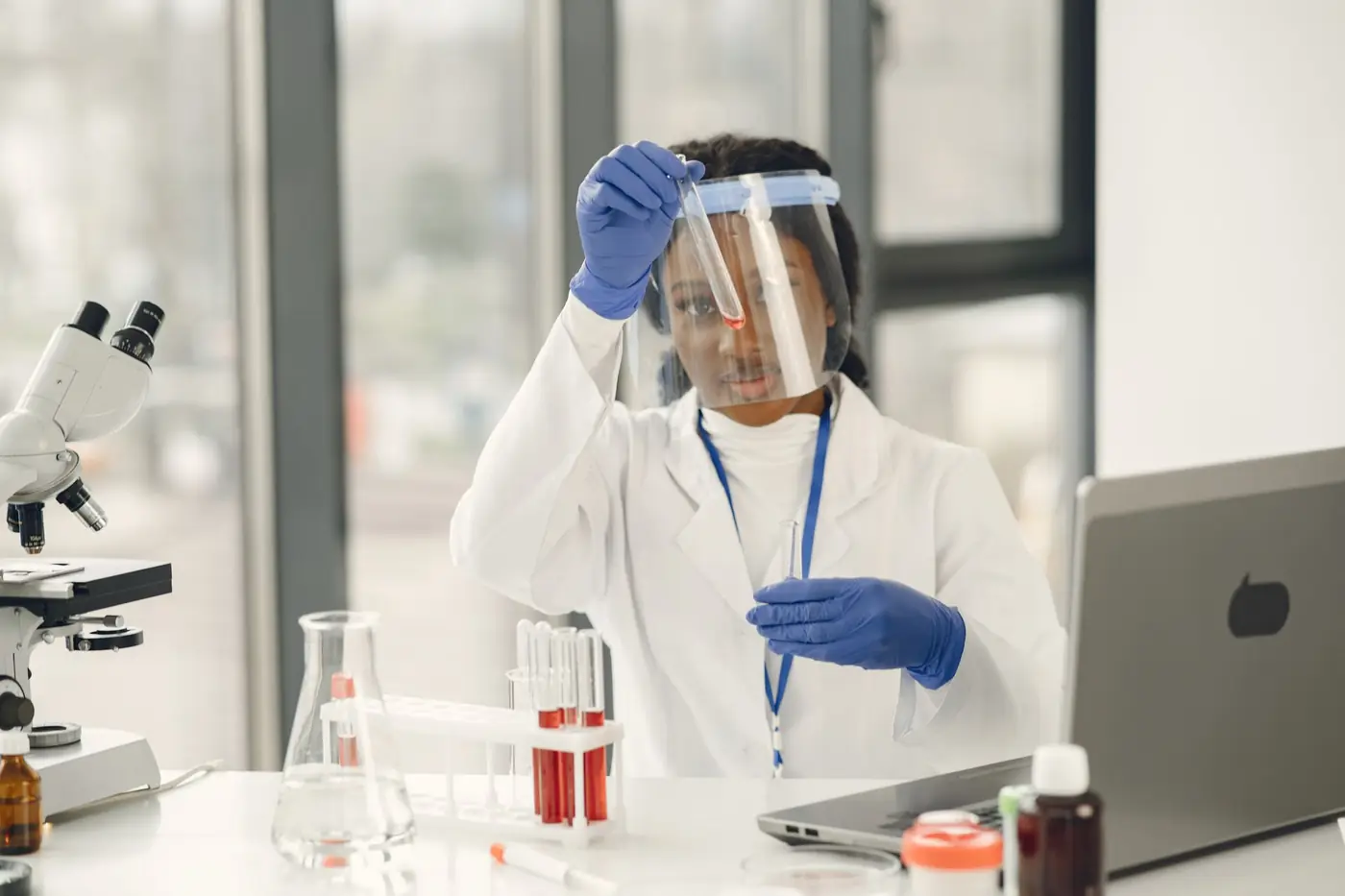 A researcher in a face shield and gloves holding a sample tube at a laboratory bench.